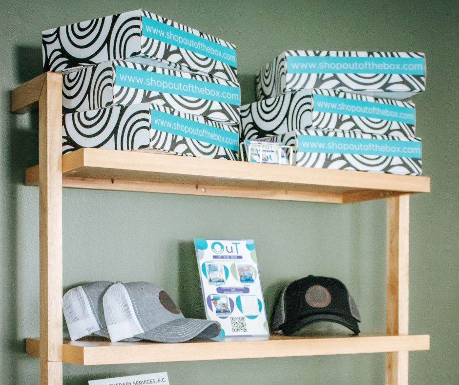 Shelves inside the Excel Therapy Services office with OuT of the Box occupational therapy boxes, caps, and a book against a green wall